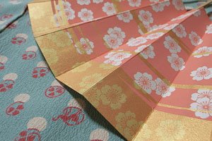 Eine nahaufnahme eines Sensu fächers mit blumen motiven. Es ist innen Pink mit weißen blüten und außen orange mit gelblichen blüten.