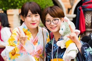 zwei frauen im Kimono. Dsie zeigen das Peace Zeichen mit den fingern. Die linke hat eine runde brille an und hält einen Grauen Teddy Bear in der Hand.